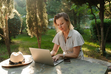 a beautiful mature woman in glasses works on a computer at a white table in nature and spends her...