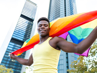 Smiling young man wearing yellow vest holding rainbow flag in front of modern buildings