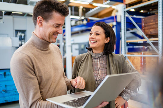 Smiling businessman holding laptop standing by businesswoman in industry