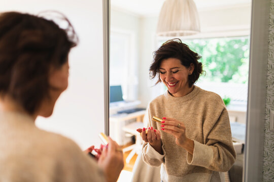 Happy Woman Holding Lipstick In Front Of Mirror At Home