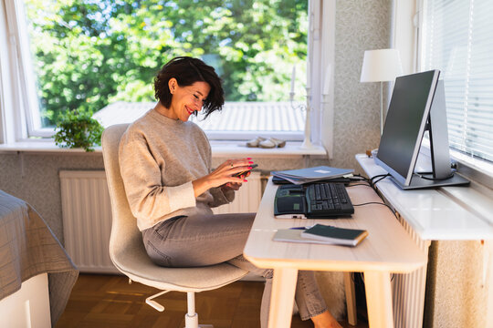 Smiling businesswoman using smart phone in front of desktop PC on table at home - Powered by Adobe