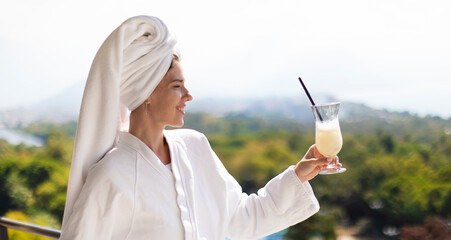 Beautiful young female wearing white hotel bathrobe enjoying vacation with cold cocktail Pina colada on a seaside balcony room apartments. Real estate advertisement. - obrazy, fototapety, plakaty
