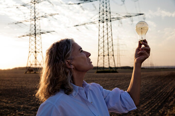 Thoughtful woman looking at light bulb in front of electricity pylons
