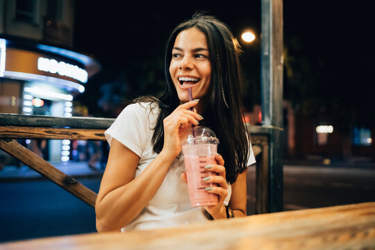 Cheerful Woman With Smoothie Sitting In Sidewalk Cafe At Night