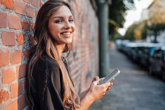 Happy young woman with mobile phone standing by brick wall