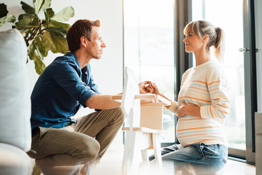 Romantic man holding hand of pregnant woman crouching near rocking horse at home