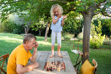 Cute girl clapping hands by grandfather playing chess on table in garden