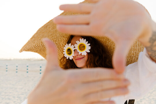 Smiling woman wearing sunflower sunglasses and oversized straw hat gesturing finger frame at beach