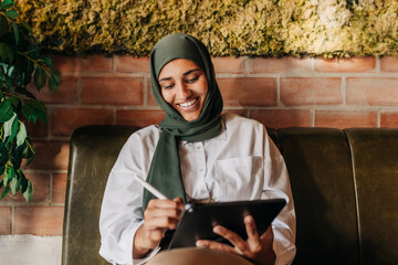 Happy woman with a hijab drawing on a digital tablet in a cafe