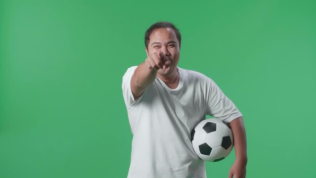 Asian Man With A Ball Pointing To Camera For Making Fun Of The Losers While Cheering Soccer On Green Screen Background
