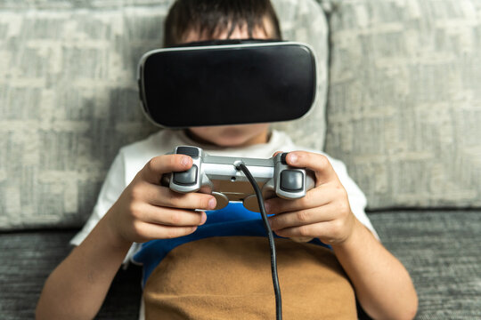Frontal image of a child lying on the sofa with virtual reality goggles connected to a game console controller playing an online game.