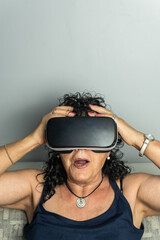 Fototapeta premium Vertical shot of curly-haired brunette senior woman wearing virtual reality goggles gaping and touching her head in amazement.