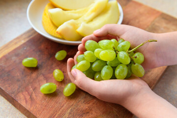 a woman's hand with green grapes, plate of melon slices on a wooden background, concept of fresh fruits and healthy food