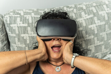 Curly-haired brunette senior woman wearing a virtual reality goggles gaping and touching her cheeks...