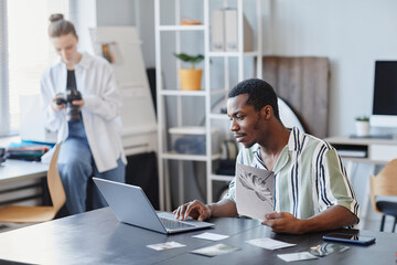 Side view portrait of black male photographer using laptop for photo editing in minimal studio setting, copy space