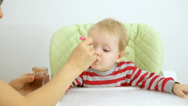 funny baby girl eats bland mashed food sitting, on high chair, mother feeds child, hand with spoon for vegetable lunch, baby weaning, first solid food for young kid.