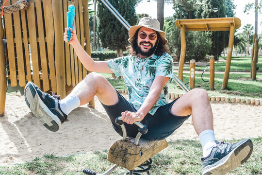 Portrait of very funny man with a lot of energy rides a wooden toy in the park holding a melted ice cream in his hand. Young adult having fun in playground wearing hat and goggles while having fun out