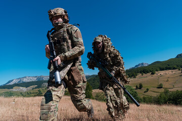 A sniper team squad of soldiers is going undercover. Sniper assistant and team leader walking and aiming in nature with yellow grass and blue sky. Tactical camouflage uniform.