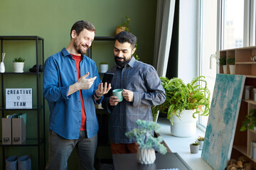 Fototapeta premium Happy young designer in smart casualwear showing his colleague new popular trends in smartphone during discussion at meeting