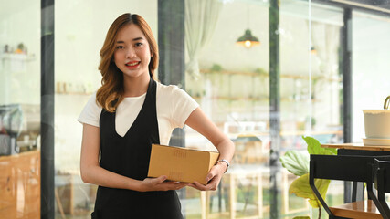 Fototapeta premium Beautiful female entrepreneur wearing apron holding cardboard and smiling to camera. Online selling, E-commerce concept