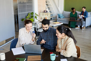 Confident man pointing at laptop screen while discussing data or video with two young female colleagues watching online training