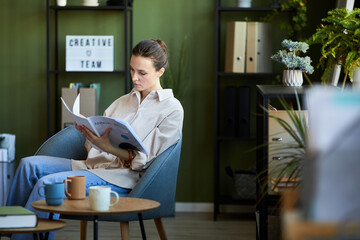 Young manager or economist in casualwear sitting in comfortable armchair and looking through manual...