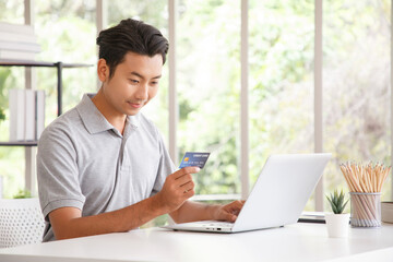 Happy cheerful smiling young adult asian man doing online shopping or e-shopping satisfied entrepreneur making online payment paying for service or goods.