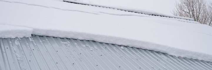 snow sliding down from roof. building construction house with metal roof covered fresh icy frozen snow and snowflakes on frosty winter day in country village suburb. snowy winter season. banner