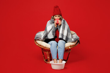 Full size young woman wear grey plaid scarf hat sit in chair soaring feet in basin drink tea hold cup isolated on plain red background Healthy lifestyle ill sick disease treatment cold season concept