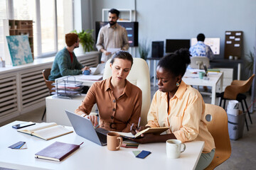 Young confident female designer explaining her colleague data while pointing at laptop screen against group of coworkers