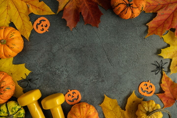 Two yellow dumbbells with Halloween decorations and small autumn pumpkins on fall leaves. Healthy fitness lifestyle composition frame. Gym workout sport training flat lay concept with copy space.
