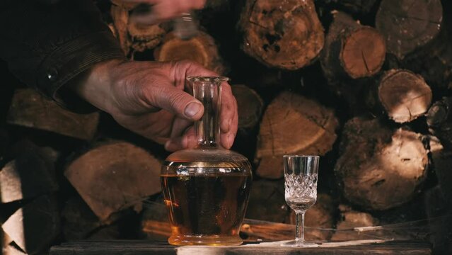 Cognac pouring in the glass from bottle. Hand puts a carafe with golden cognac and pouring into the glass, wooden table near logs. Strong drink - brandy or whiskey with ice. Alcohol amber drink
