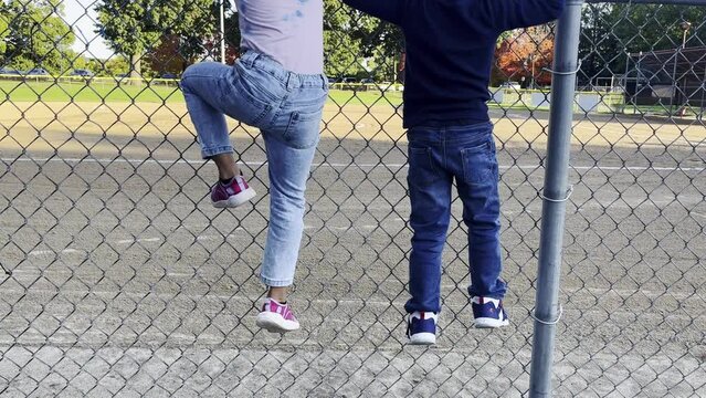 Two toddlers climbing chain link fence at softball field on a Sunny day, close up static shot