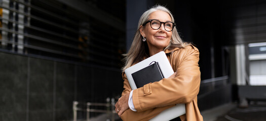 portrait of an elderly businesswoman with a laptop in glasses outside the office, strong and...