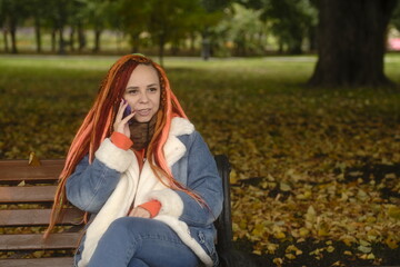 Attractive young woman in warm clothes sitting on bench and talking on mobile phone in park