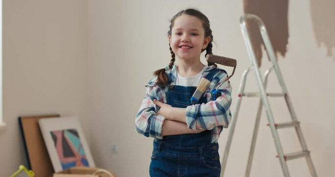 Beautiful girl with pigtails stands in room, puts hands together, looks at the camera and smiles. She is wearing checkered shirt and denim overalls. Girl is holding roller and brush. Behind is ladder.