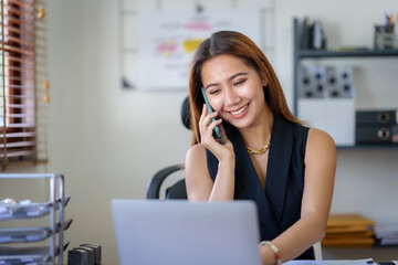 Obraz premium Asian businesswoman sitting with her laptop happily chatting on the phone with a customer explaining in detail the company's products in the office.