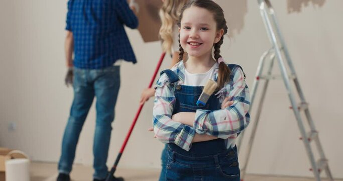 Cheerful girl with pigtails stands in the middle of room, puts hands together, looks at the camera and smiles. She is wearing checkered shirt and denim overalls. Dad and mom paint wall in brown.