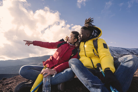 Multiethnic Couple Of Young People Relaxing On The Top Of The Mountain Sitting On The Floor And Pointing Fingers Looking Away - Wanderlust Hiker People And Diverse Couple Lifestyle Concept