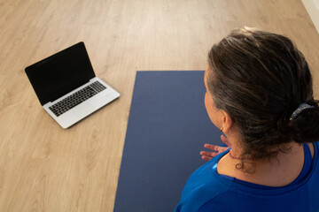 The yoga teacher, dressed in blue, is sitting on a mat looking at her computer to start the online class.