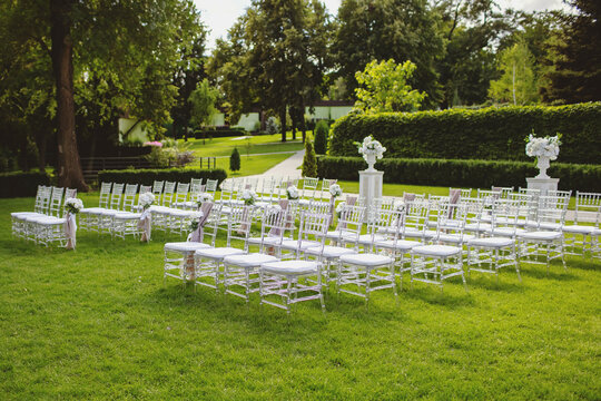 White chairs on a green lawn for a wedding ceremony are waiting for guests. Beautifully decorated seating areas in the form of chairs with flowers and fabrics. Preparing for the wedding ceremony