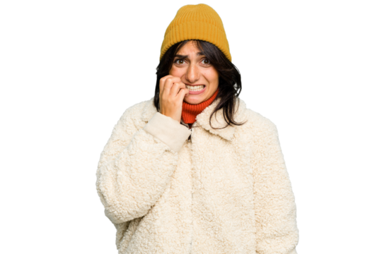 Young Indian woman wearing winter jacket and a wool cap isolated biting fingernails, nervous and very anxious.