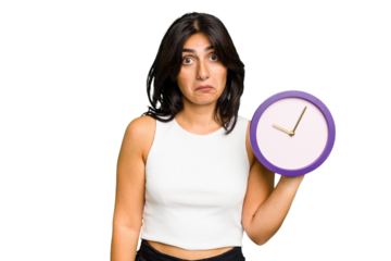 Young Indian woman holding a clock isolated shrugs shoulders and open eyes confused.