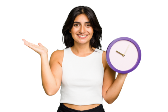 Young Indian woman holding a clock isolated showing a copy space on a palm and holding another hand on waist.