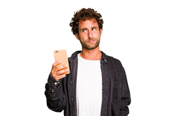Young caucasian man using mobile phone isolated confused, feels doubtful and unsure. - obrazy, fototapety, plakaty