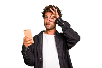 Young caucasian man using mobile phone isolated excited keeping ok gesture on eye.