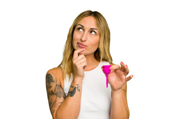 Young caucasian woman holding menstrual cup isolated on green chroma background looking sideways with doubtful and skeptical expression.