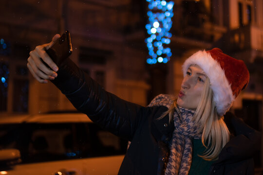 Happy young woman in a Santa Claus hat taking a selfie against the background of the Christmas lights of the night city. Concept of holidays and people