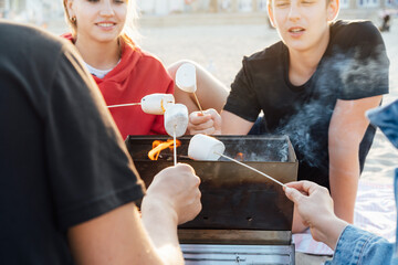 Group Of Young Friends Toasting Marshmallows Around Fire On the beach. Active Teenagers outdoor activity. Close up grilled sweets on the fire. Sunset time. Selective focus.