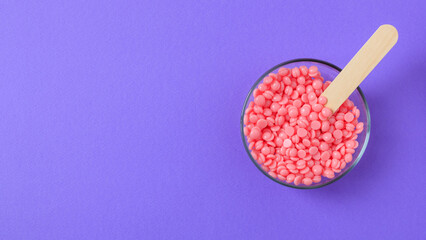Pink granules of wax for depilation in a glass bowl on a lilac background. Place for text. Epilation, depilation, waxing, unwanted hair removal procedure. - obrazy, fototapety, plakaty